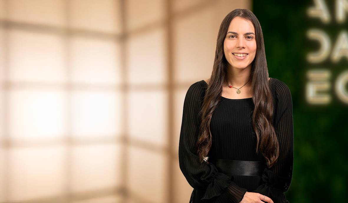 Una mujer sonriente con cabello largo y suelto, vestida con un atuendo negro, está de pie frente a un fondo iluminado.
