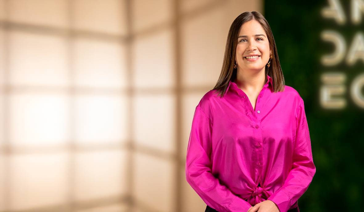 Una mujer sonriente viste una camisa rosa y se encuentra en un entorno iluminado con fondo desenfocado.