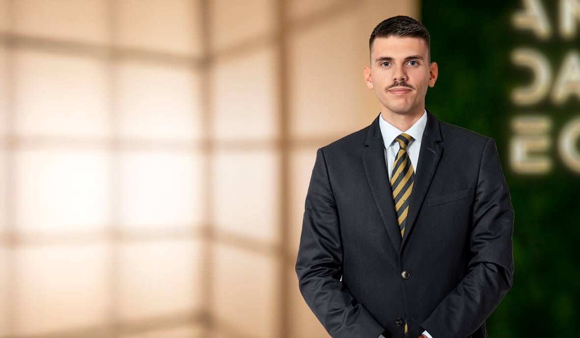 Un hombre de pie con un traje formal y una corbata en un fondo desenfocado.