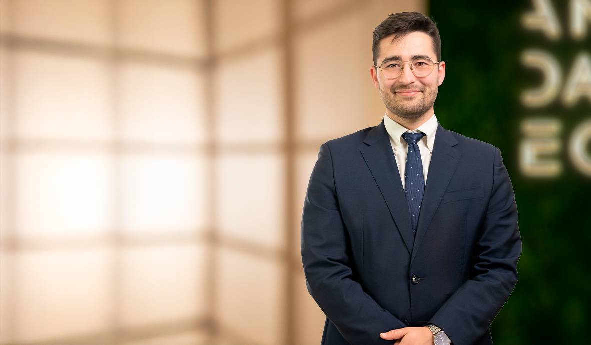 Un hombre joven sonriente vestido con traje en un fondo desenfocado.