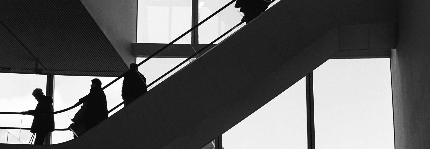 Una escena en blanco y negro que muestra a varias personas subiendo una escalera en un edificio moderno con grandes ventanales.