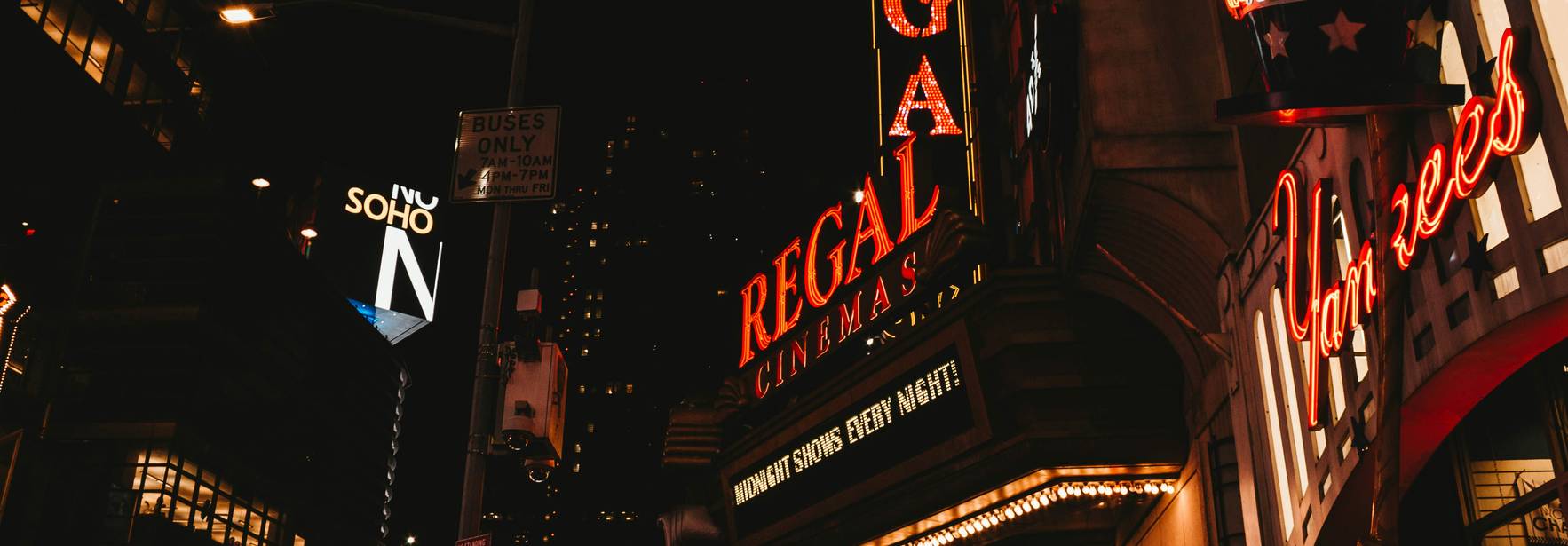 La imagen muestra un cine Regal iluminado por neón en una concurrida calle de la ciudad durante la noche.