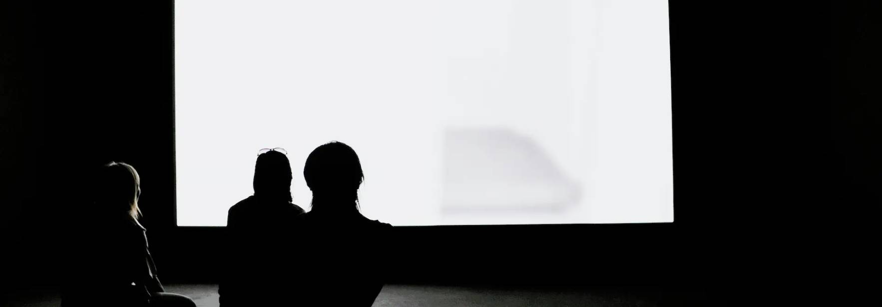 Silhouette de tres personas observando una pantalla blanca en un ambiente oscuro.
