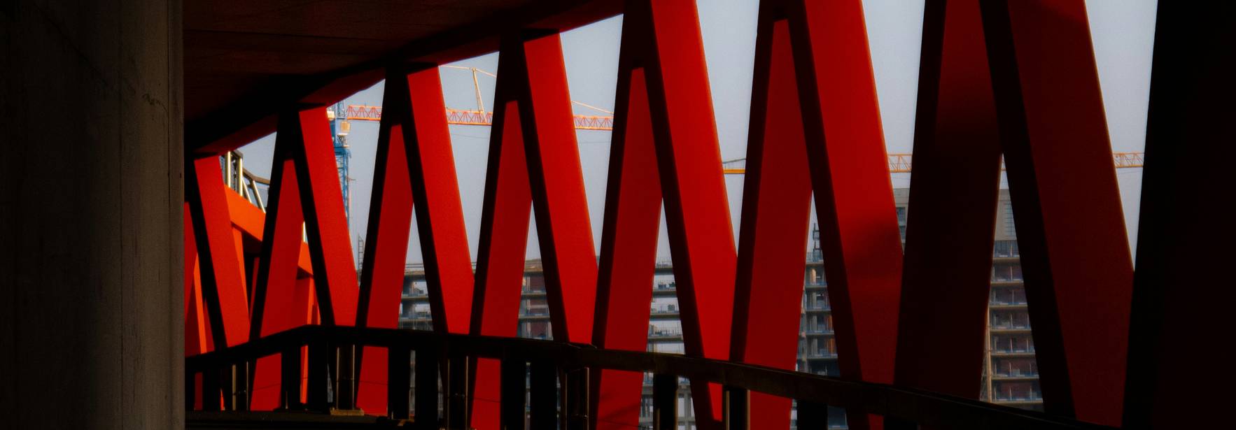 Una estructura arquitectónica con soportes rojos y un pasillo oscuro que muestra edificios en construcción al fondo.