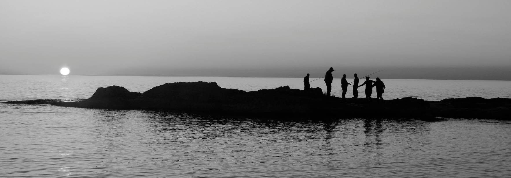 Siluetas de personas en una roca al atardecer, con el sol poniéndose en el horizonte.