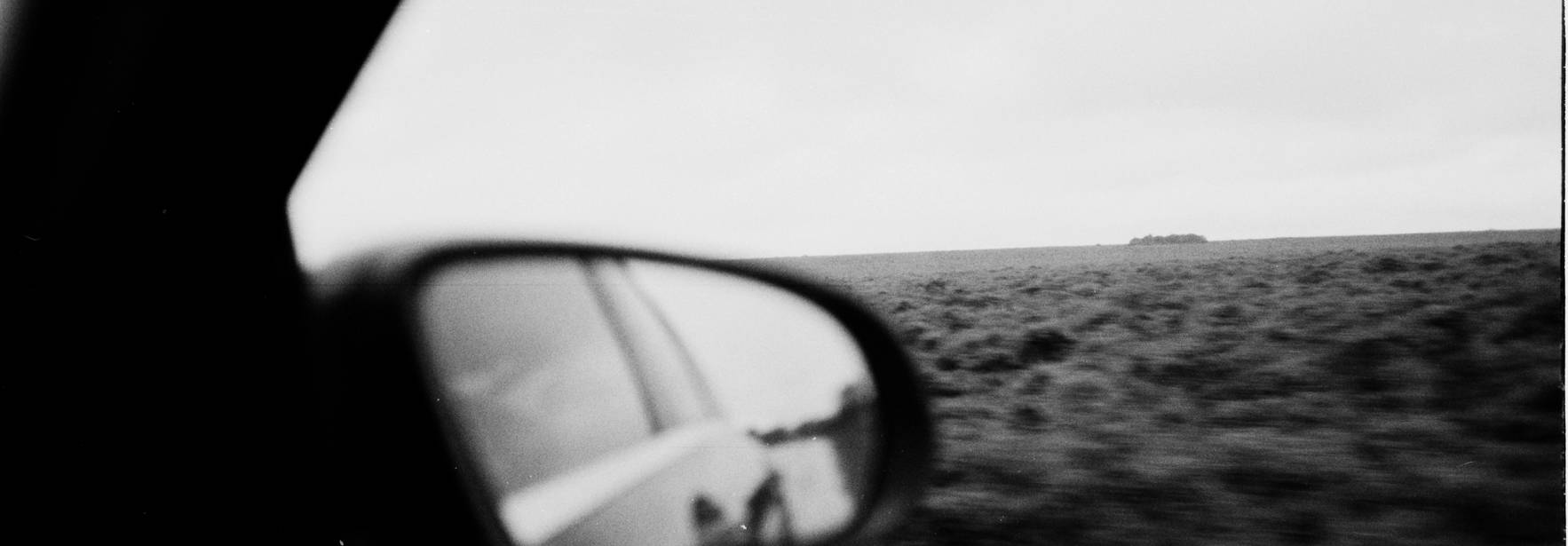 Una vista en blanco y negro desde el interior de un coche, mostrando el paisaje del exterior reflejado en el espejo lateral.