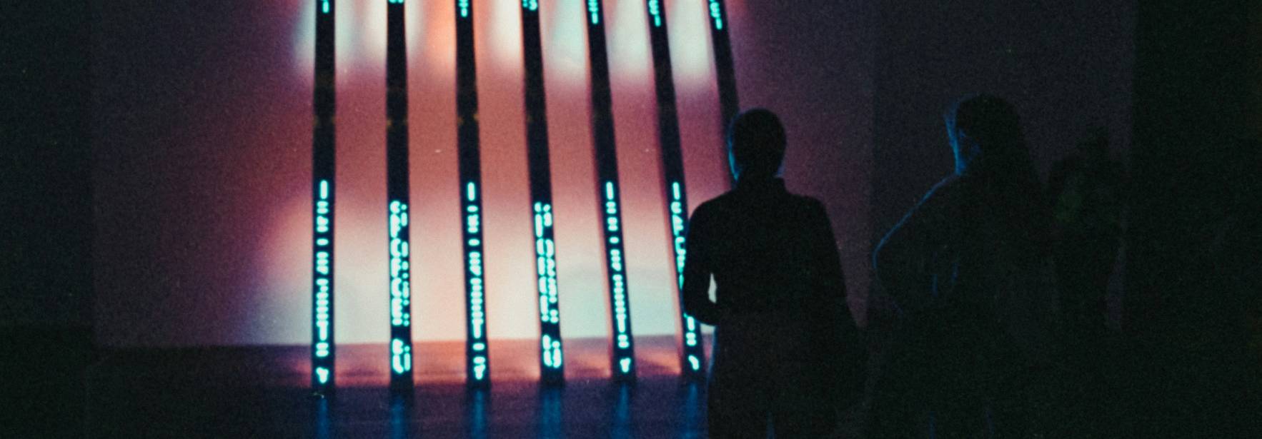 La imagen muestra a dos personas observando una instalación artística iluminada con luces azules y rojas en una sala oscura.