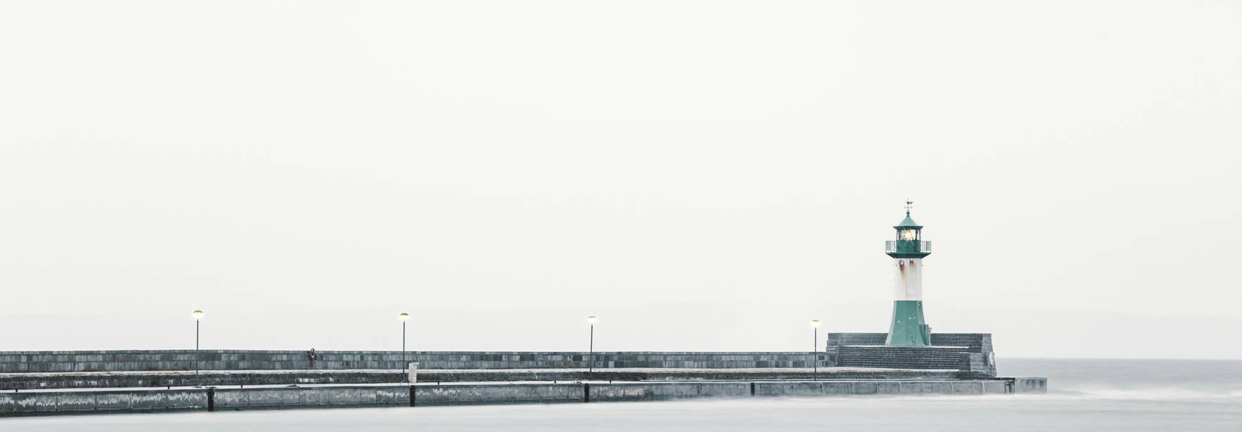 Un faro solitario se encuentra en un muelle sobre un mar tranquilo y grisáceo.