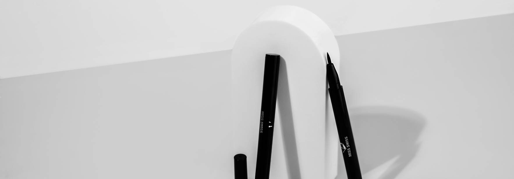 Una composición artística que presenta un soporte blanco en forma de letra y varios marcadores negros en una mesa clara.