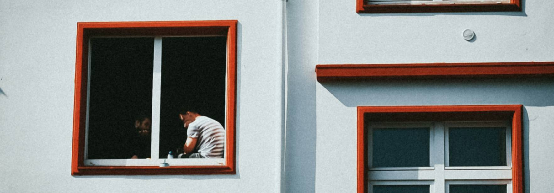 Una persona observa desde una ventana con marcos rojos en un edificio blanco.