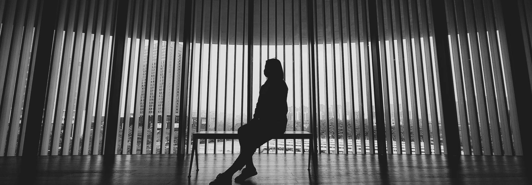 Una figura humana se sienta en un banco frente a una serie de lamas verticales en un entorno urbano.