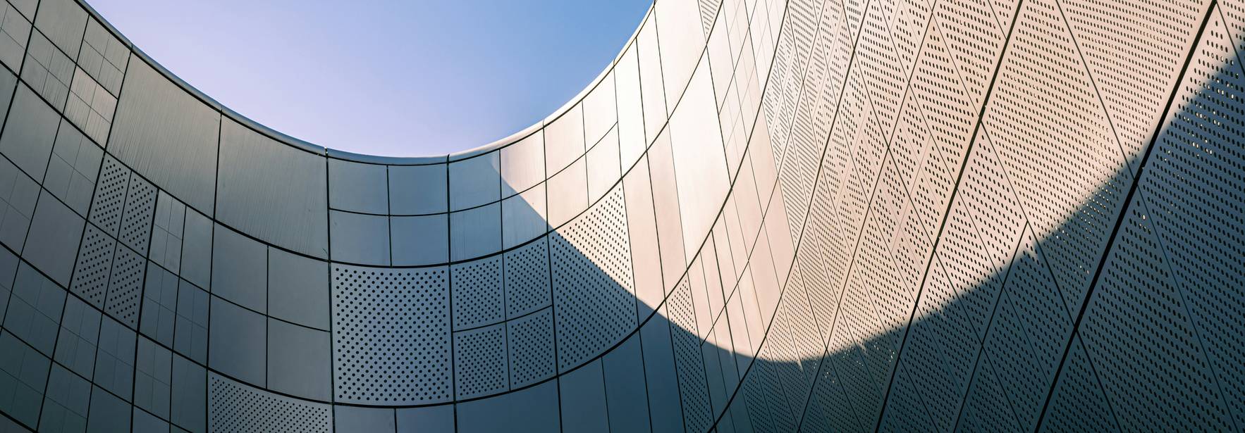La imagen muestra una vista desde abajo de una estructura arquitectónica moderna con paredes curvas y un cielo azul de fondo.