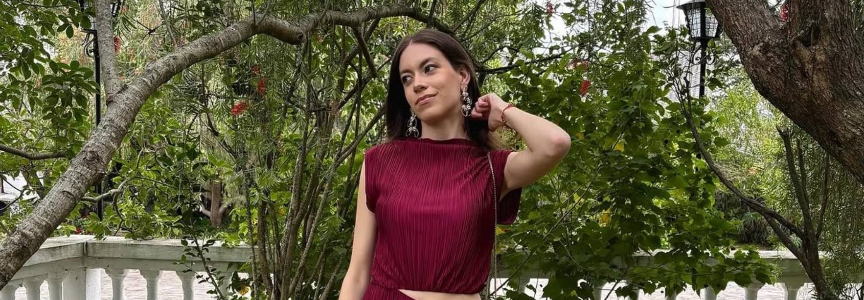 Una mujer posa con un vestido corto de color burdeos en un entorno natural con árboles y un fondo de columnas blancas.