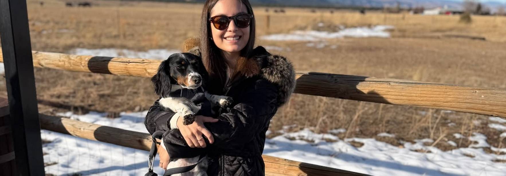 Una joven sostiene un perro en un paisaje montañoso con cielo despejado.