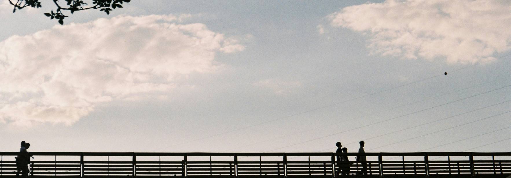 La imagen muestra un puente con siluetas de personas caminando bajo un cielo parcialmente nublado.
