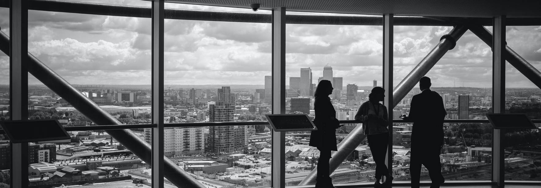 Tres personas de pie frente a un ventanal observando la vista de una ciudad desde un edificio alto.