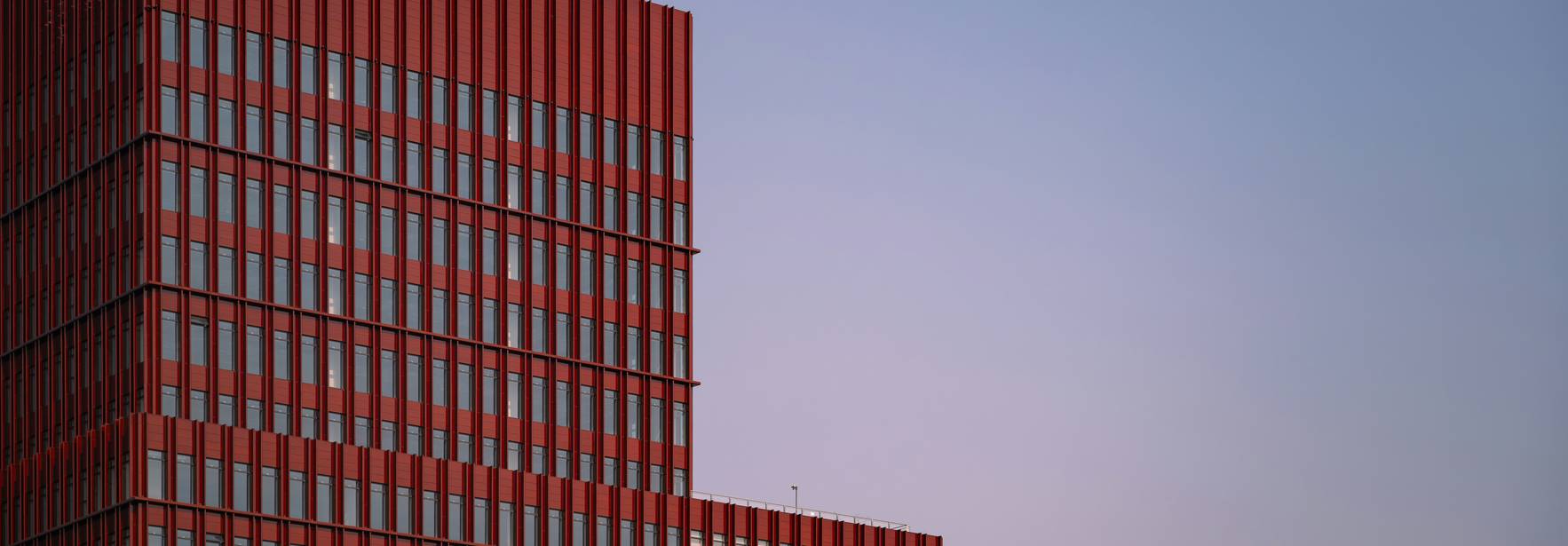 Un edificio moderno de color rojo con ventanales, destacándose en un cielo suave al atardecer.
