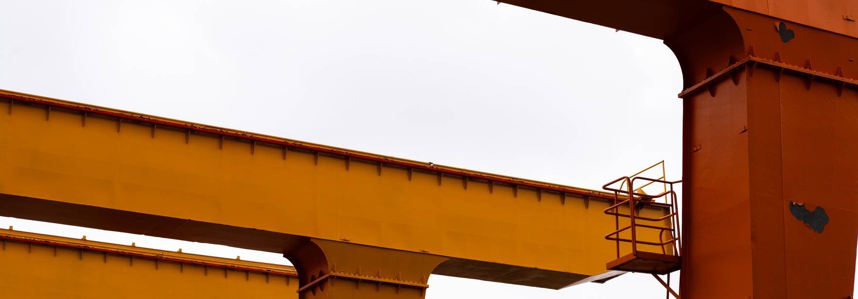 La imagen muestra grúas de carga de color naranja y amarillo con un cielo gris al fondo.