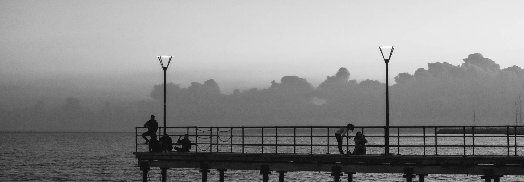 Una escena en blanco y negro de un muelle al atardecer con personas sentadas y la calma del mar.