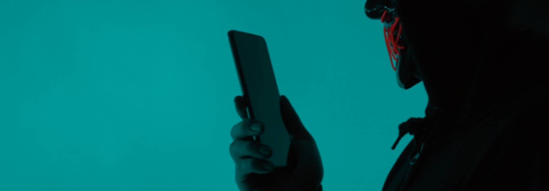 Una figura con capucha sostiene un teléfono móvil frente a un fondo azul.