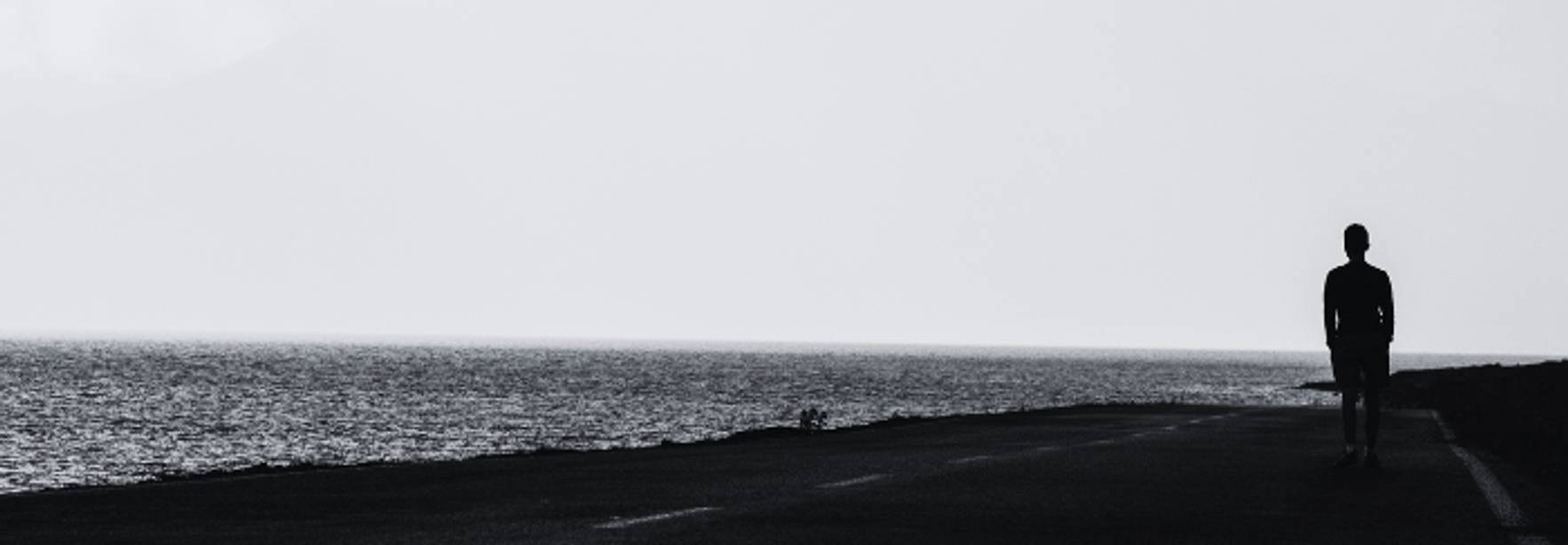 Una persona camina sola junto a una carretera frente al mar en una escena en blanco y negro.