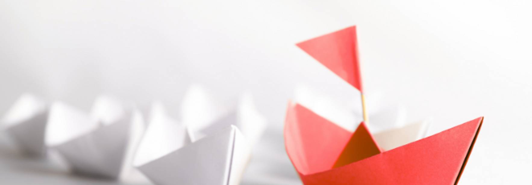 Una serie de barcos de papel, destacando un barco rojo en primer plano frente a varios barcos blancos.