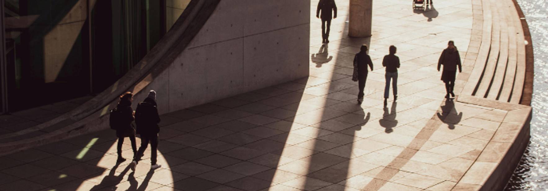 Varias personas caminan por una pasarela con sombras marcadas por la luz del sol.