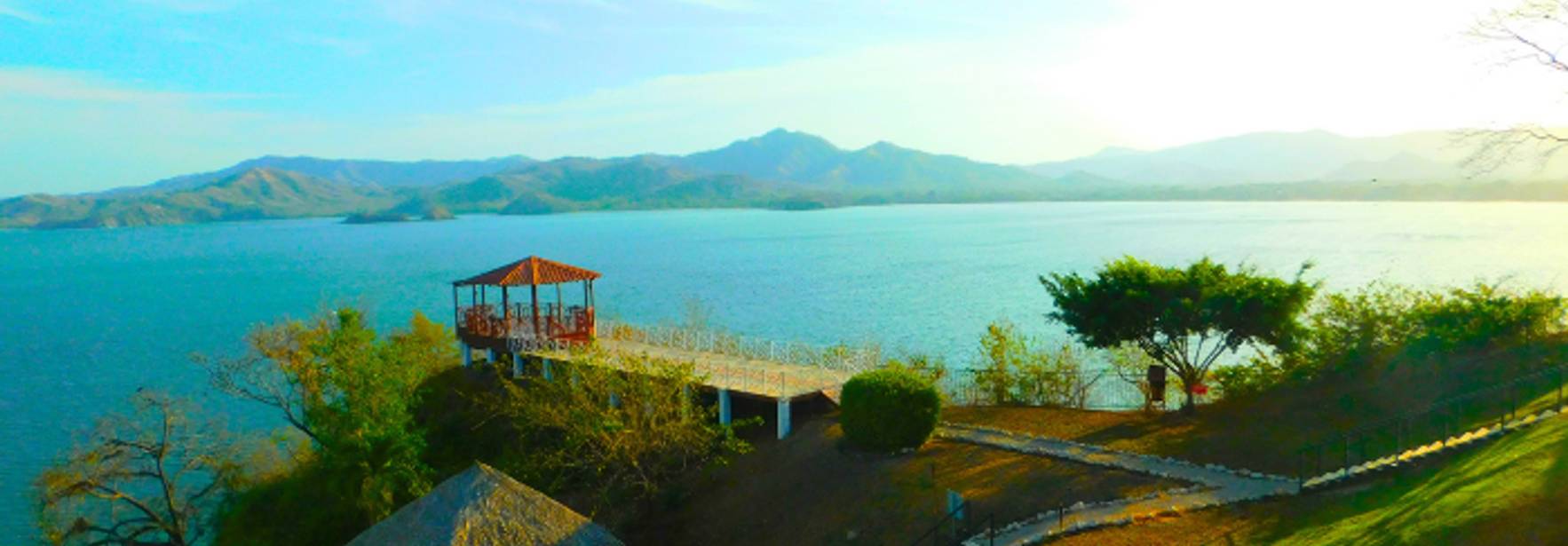 Vista panorámica de un lago rodeado de montañas y vegetación, con un muelle y una construcción a la orilla.