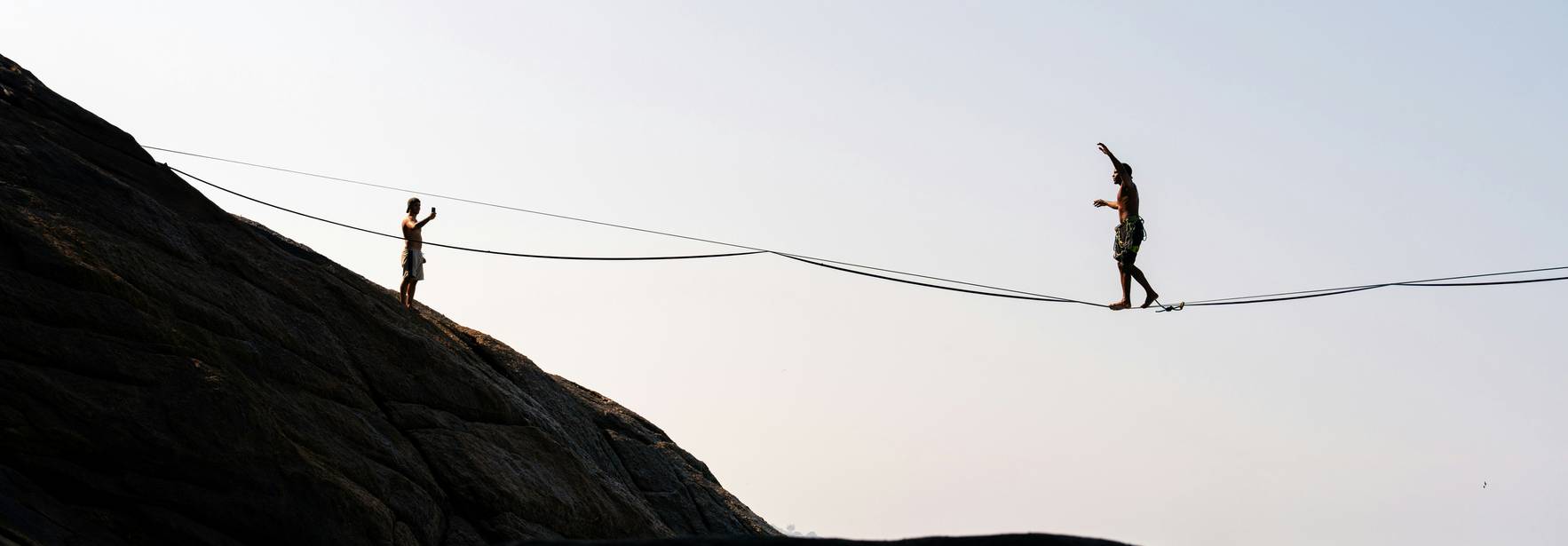 Dos personas caminan sobre una cuerda floja entre rocas cerca del mar.