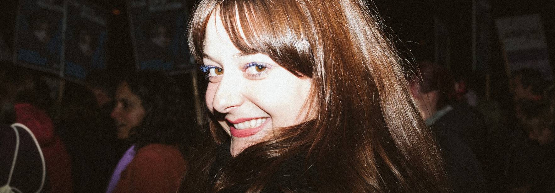 Una mujer sonriente se encuentra en un entorno de evento con personas al fondo.