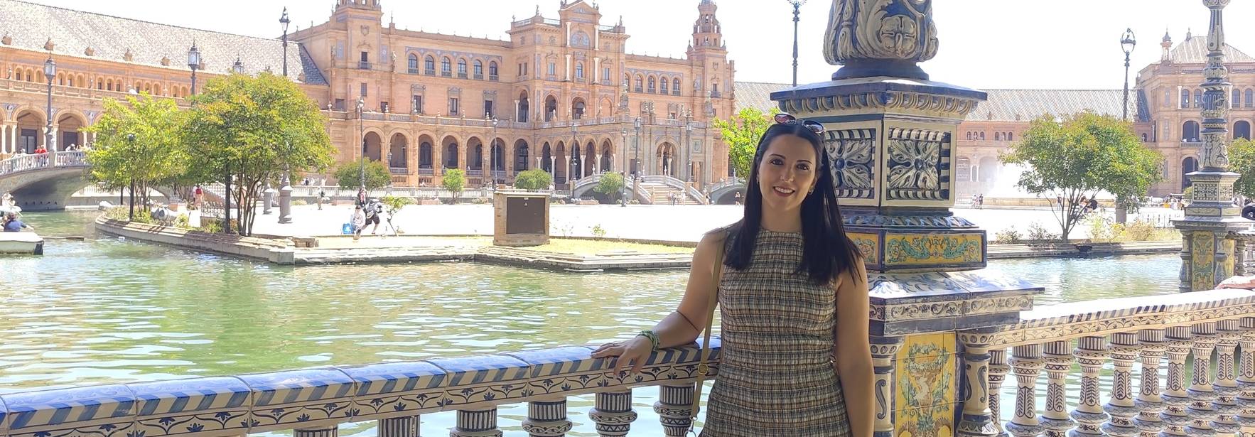 Una persona posa junto a una barandilla decorada con mosaicos, con un fondo de un edificio emblemático y un estanque.