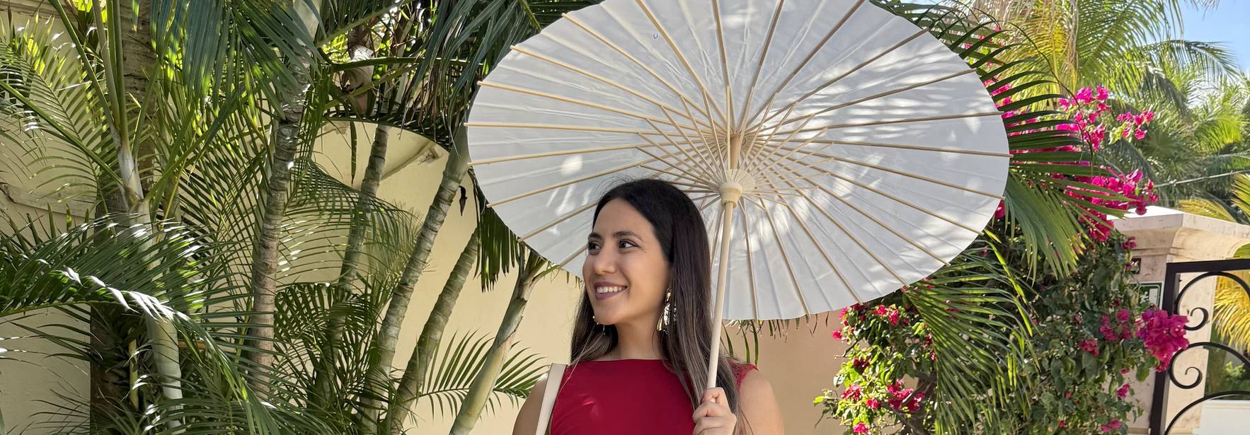Una mujer sonriente sostiene un paraguas sombrilla mientras está rodeada de plantas tropicales.