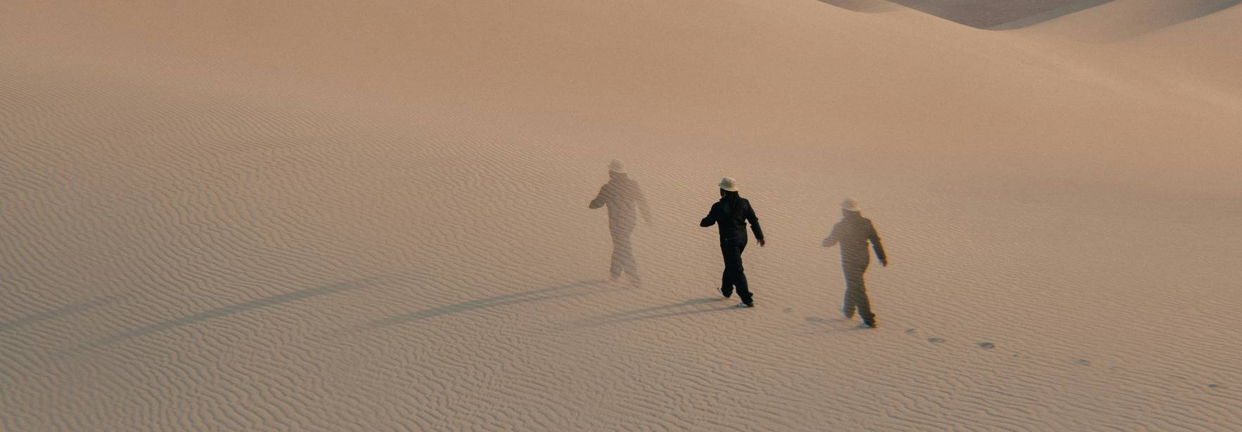 Tres personas caminan a través de un desierto de arena con oleadas suaves en el fondo.