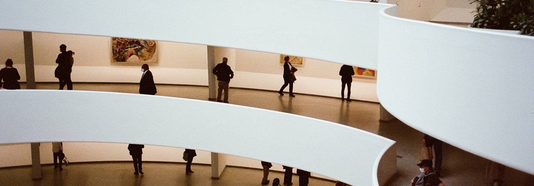 La imagen muestra un interior moderno de un museo con varias personas caminando y obras de arte visibles en las paredes.