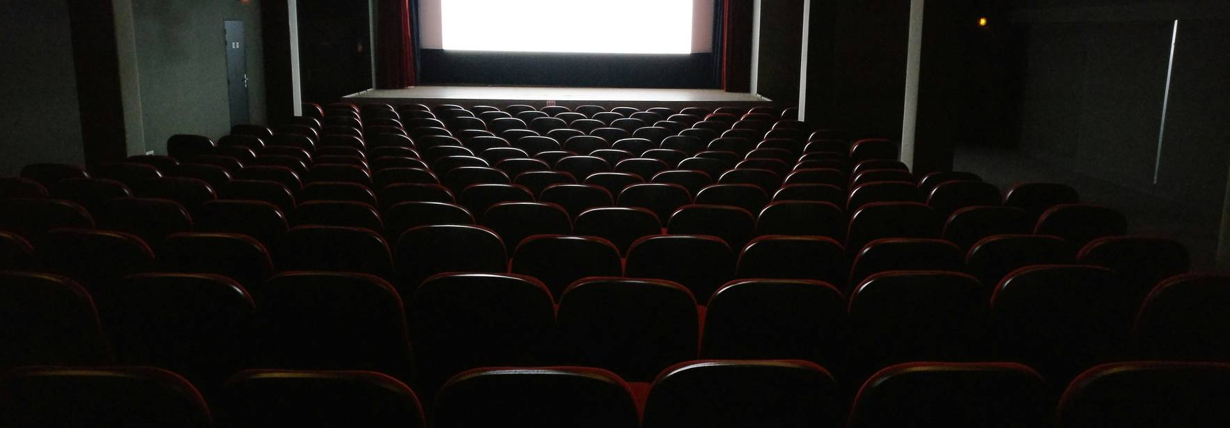 Una sala de cine vacía con asientos en filas y una pantalla iluminada al fondo.