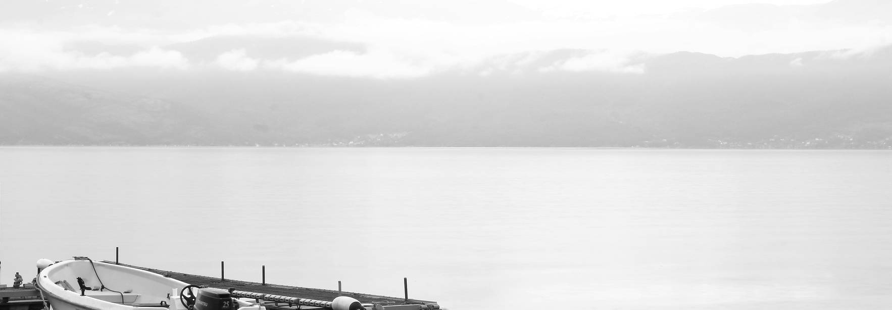 Una embarcación amarrada en un muelle tranquilo junto a un lago en un atmósfera brumosa y en blanco y negro.