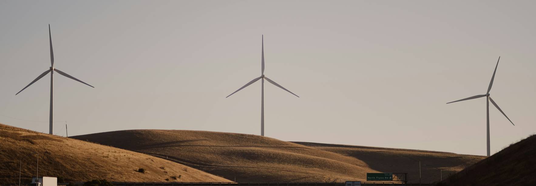 Una carretera se extiende frente a colinas con aerogeneradores altos al fondo bajo un cielo despejado.