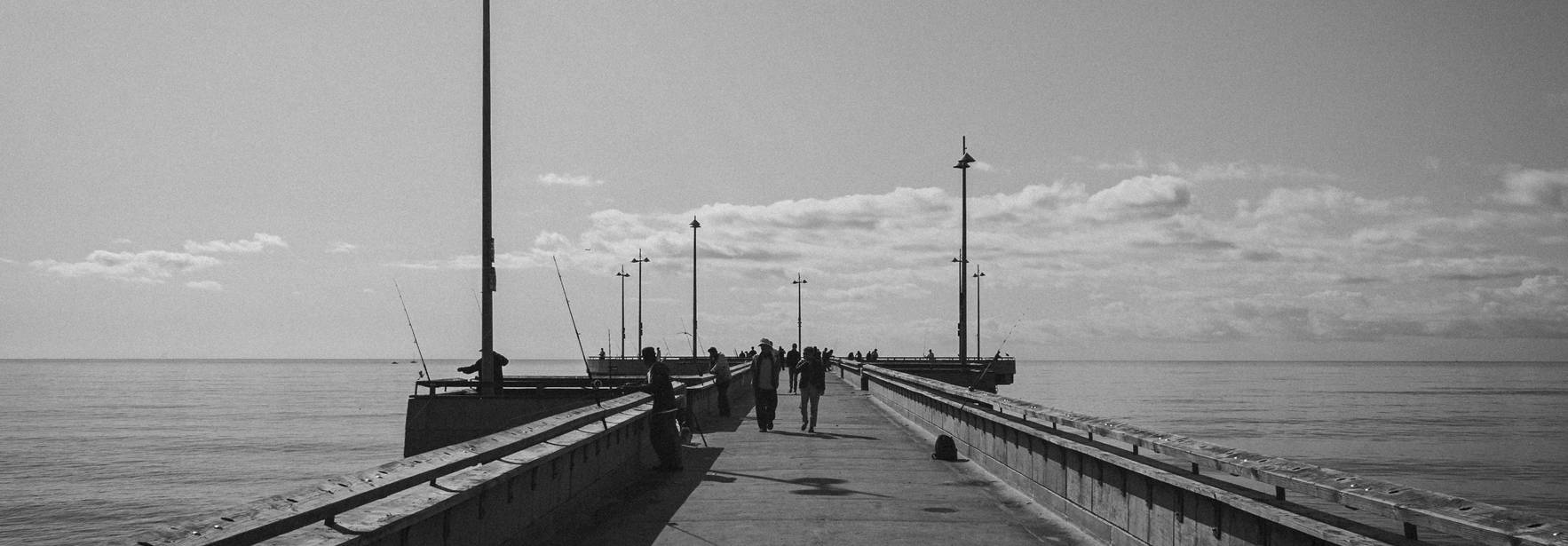 Un muelle en blanco y negro, con personas caminando y faroles a lo largo del camino.