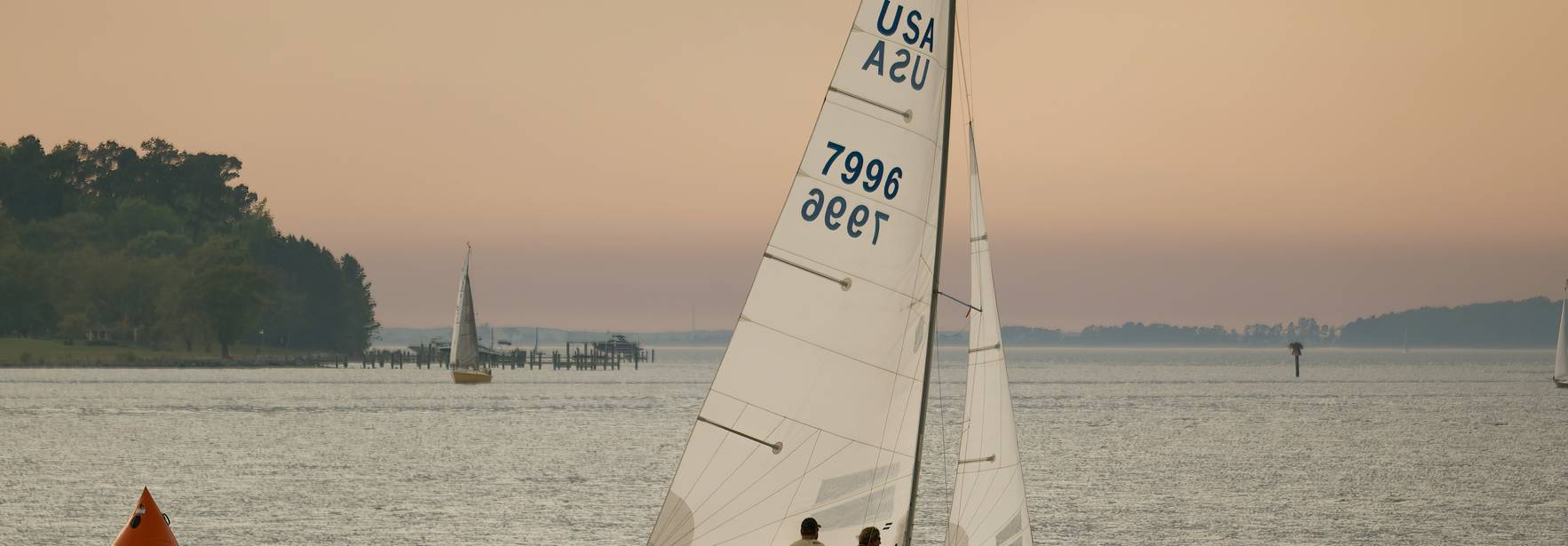 Un par de personas navegan en un velero sobre un lago al atardecer.