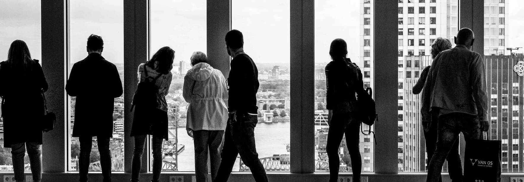 Un grupo de personas observa una vista urbana desde una gran ventana en blanco y negro.