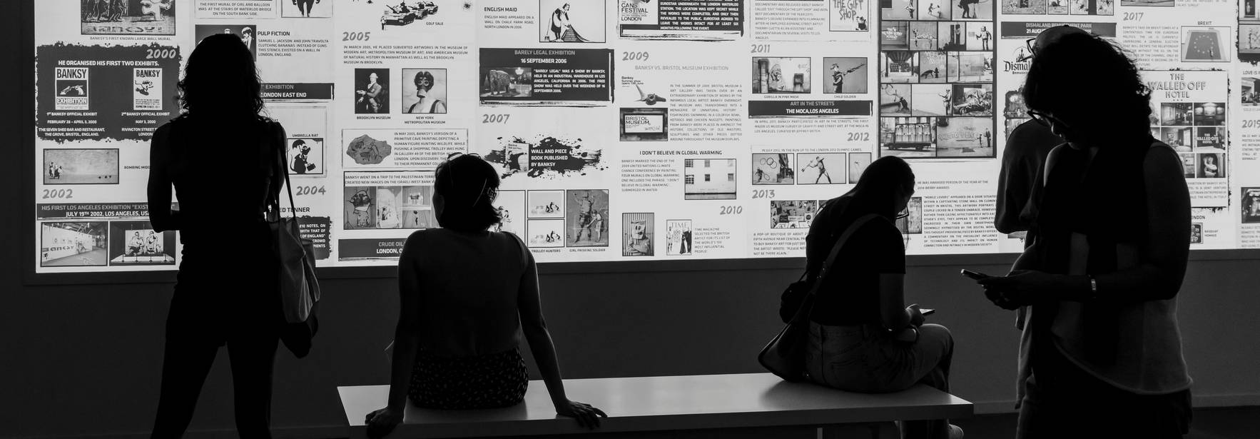Una exposición en blanco y negro donde se pueden ver personas sentadas y de pie frente a una serie de paneles llenos de imágenes y textos.