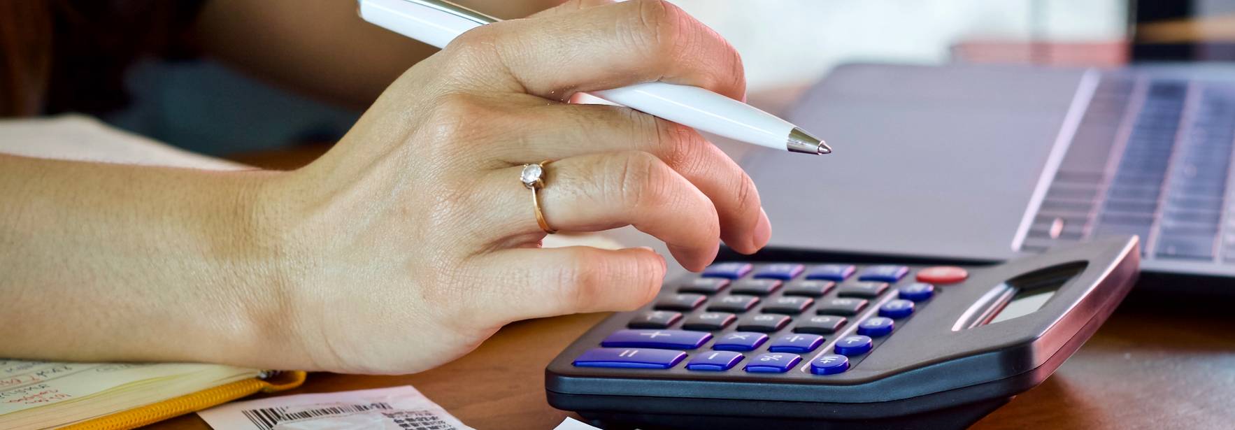 Una mano utiliza una calculadora mientras hay recibos y una laptop en la mesa.