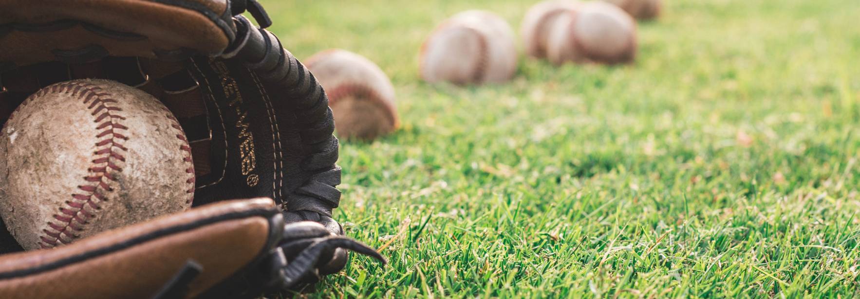 Un guante de béisbol con una pelota, situado sobre un campo de hierba, junto a varias pelotas más en el suelo.