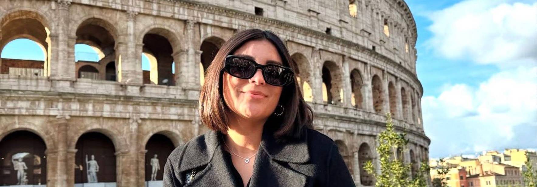 Una persona sonriente posa frente al Coliseo en Roma bajo un cielo azul.