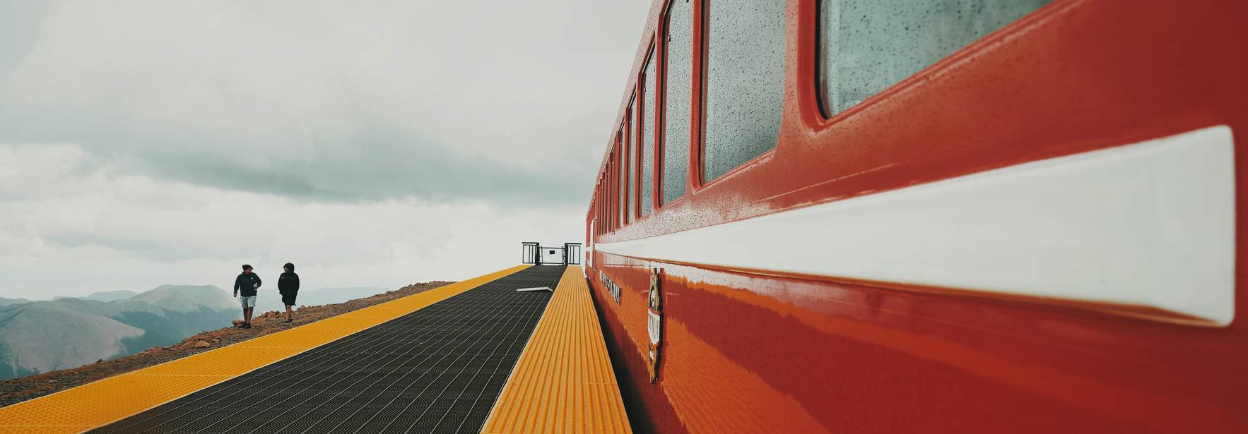 Una plataforma de tren con un tren rojo y personas caminando en un paisaje montañoso.
