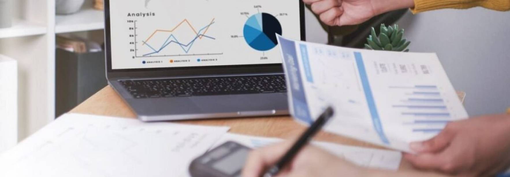 Una persona señala gráficos en una computadora portátil mientras otra escribe en documentos sobre una mesa.