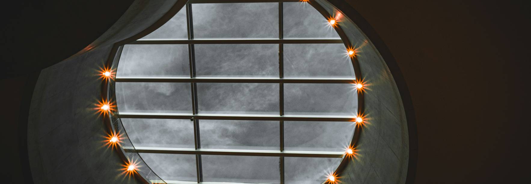 La imagen muestra una claraboya circular iluminada por luces alrededor, con nubes grises visibles a través del cristal.