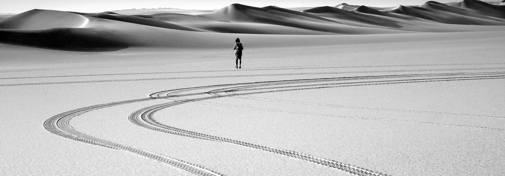 Una persona solitaria se encuentra en un vasto desierto, rodeada de ondulantes dunas de arena con huellas de neumáticos marcadas en el suelo.