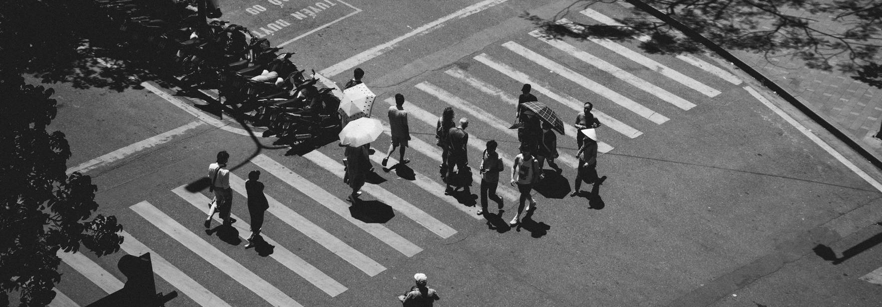 Una imagen en blanco y negro que muestra un grupo de personas cruzando una calle con un paraguas en una zona urbana.