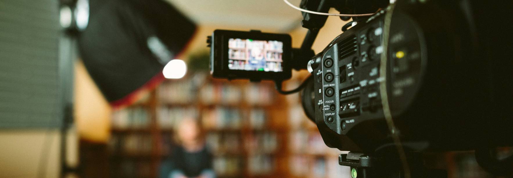 Una cámara de video en primer plano enfocando a una persona sentada en un entorno de estudio con estanterías de libros de fondo.